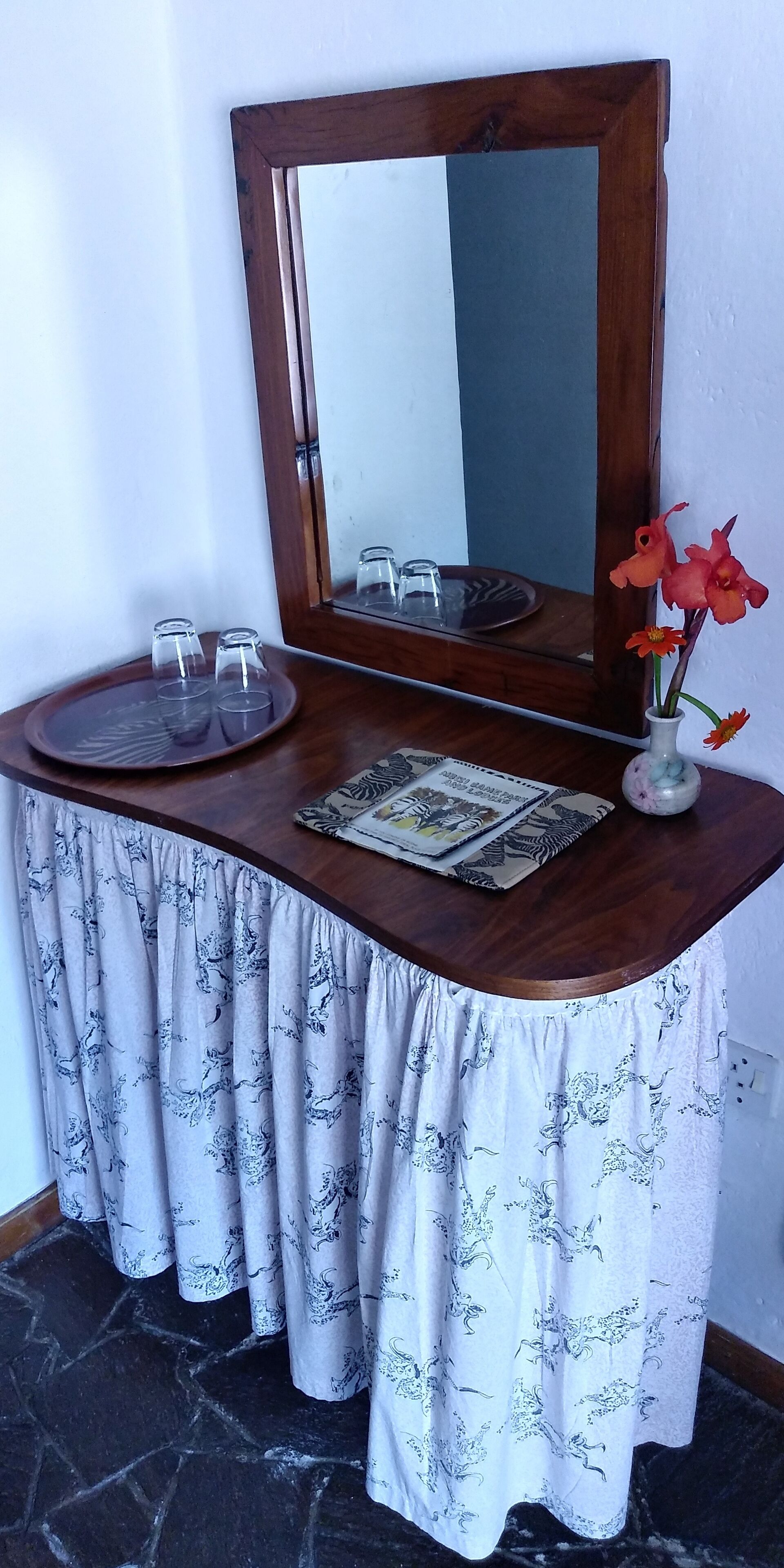 A corner table with a mirror, a tray with glasses, a vase with flowers, and patterned curtains.