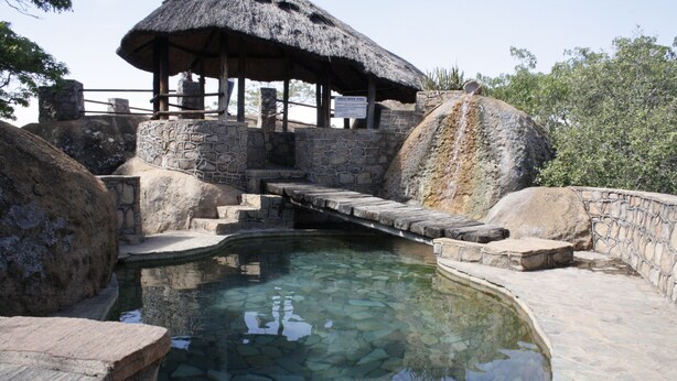 A stone bridge over a clear pool, surrounded by large rocks and a thatched-roof structure. - Ruwa River Herbal Clinic & All-Inclusive Oasis Spa Resort (Harare)