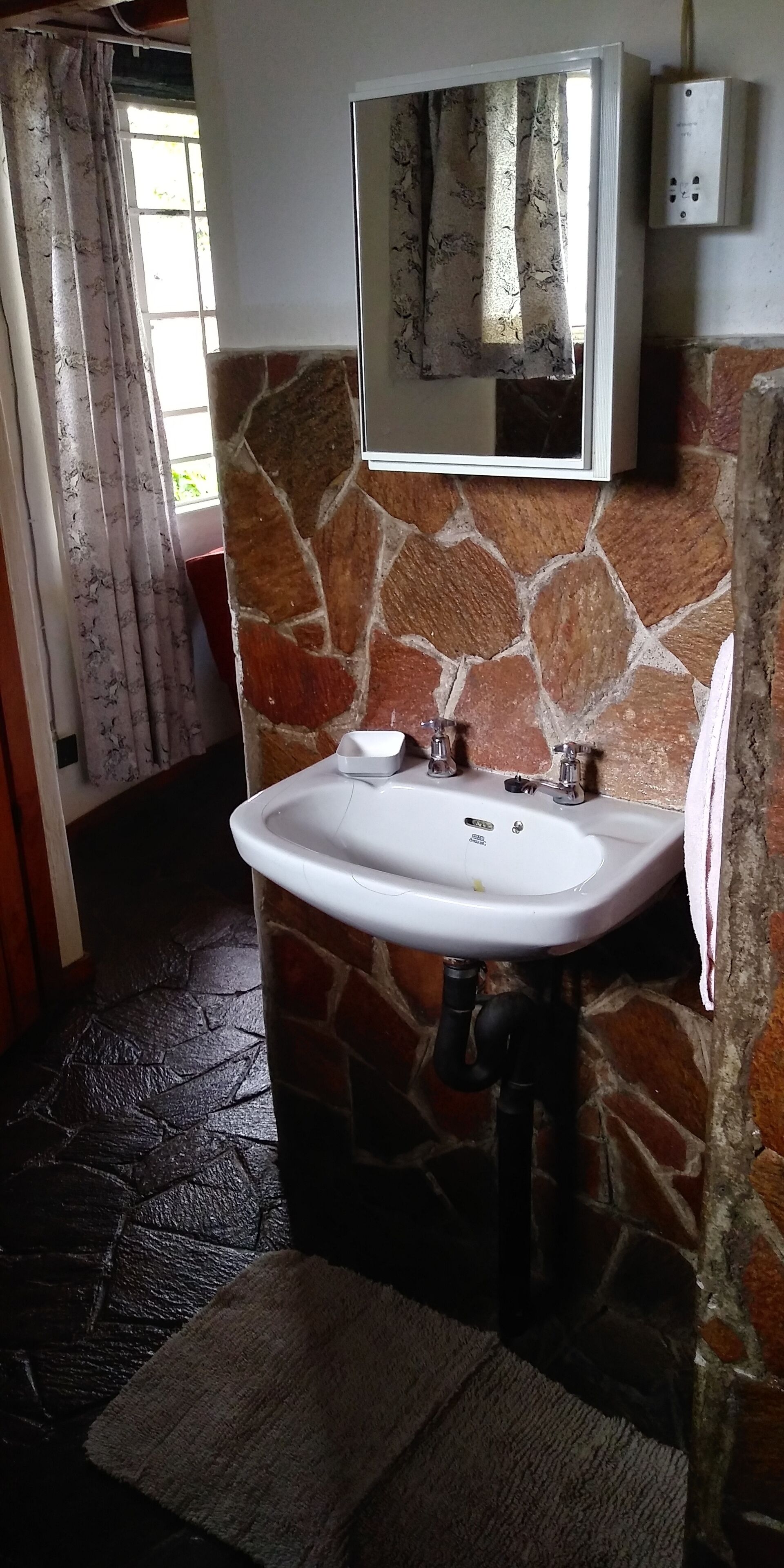 Un baño con pared de piedra, un lavamanos blanco, un espejo y una ventana con cortinas.
