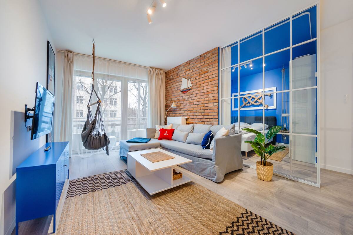 Una sala de estar moderna con sofá, mesa de centro y sillón colgante. Hay un televisor montado en la pared, un ventanal grande y una pared decorada con ladrillos.