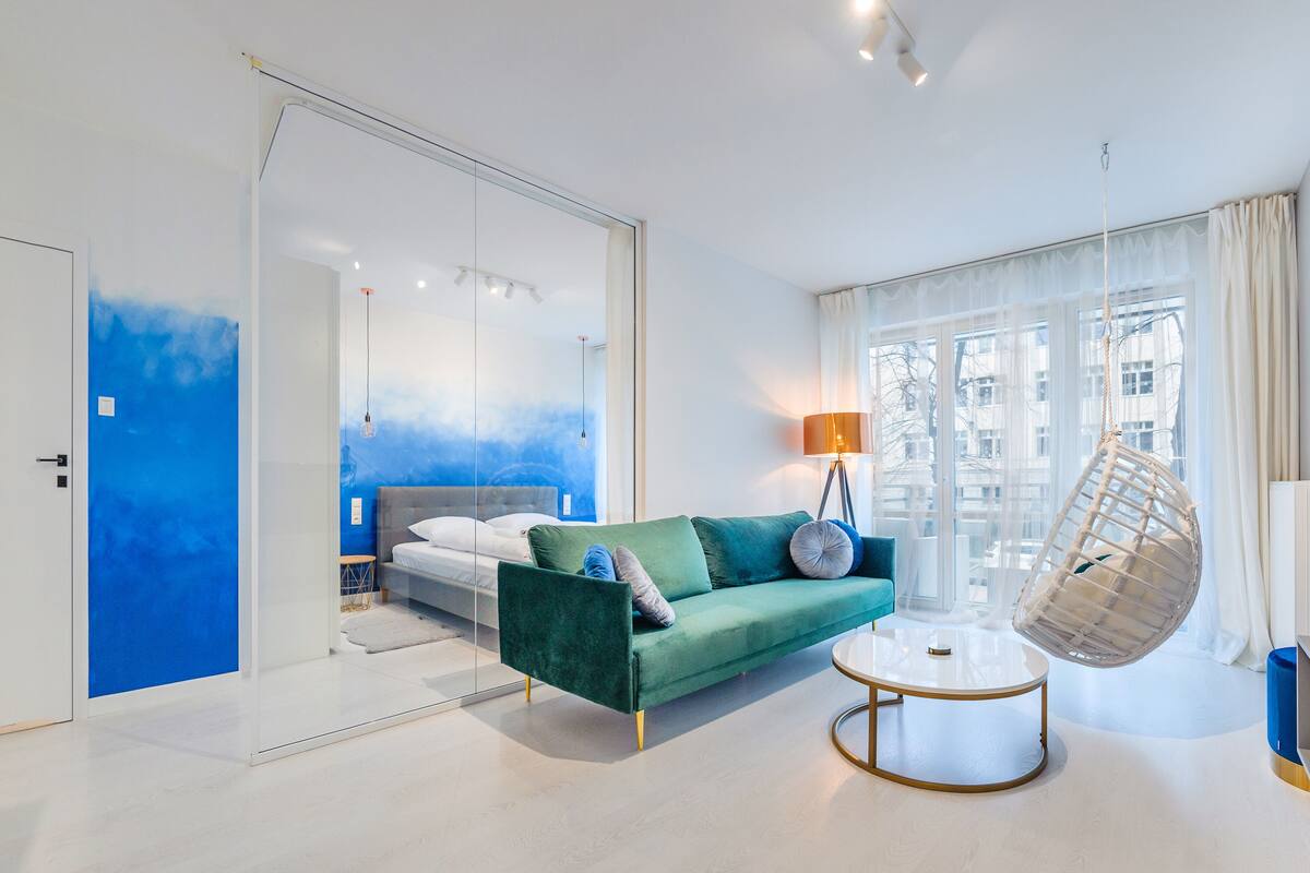 Una habitación de hotel moderna con un sofá verde, un sillón colgante, una cama con pared azul y ventanas amplias.