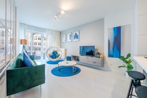 Ein modernes Wohnzimmer mit einem grünen Sofa, einem runden Couchtisch, einer weißen TV-Konsole und einem großen Fenster mit Vorhängen.