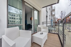Ein Balkon mit weißen Korbmöbeln, einer Glasreling und Blick auf Gebäude und Bäume.
