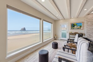 A room with large windows overlooking a beach, a wooden ceiling, and a seating area with a table.
