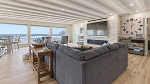 Ein großzügiger Wohnbereich mit einer großen grauen Couch, einem Holztisch und einem Flachbildfernseher, der an einer weißen Ziegelwand befestigt ist. Große Fenster bieten Blick auf den Ozean.