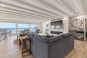 A modern living room with a large gray sectional sofa, a wooden coffee table, and a flat-screen TV mounted on a white brick wall. There are large windows offering a view of the ocean.
