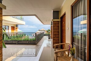 Un couloir d’hôtel donnant sur la mer, un balcon avec une balustrade et une chaise en bois sur laquelle est posé un vase de fleurs.