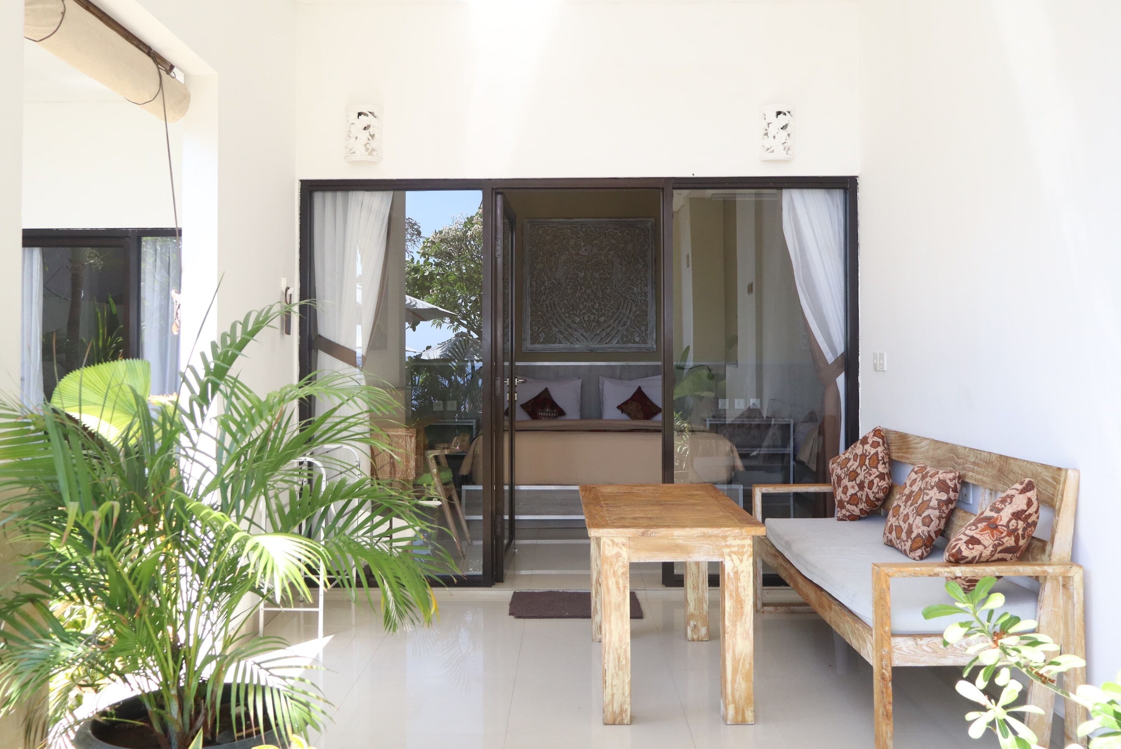A room with a wooden table and sofa, a large potted plant, and a sliding glass door leading to a balcony.