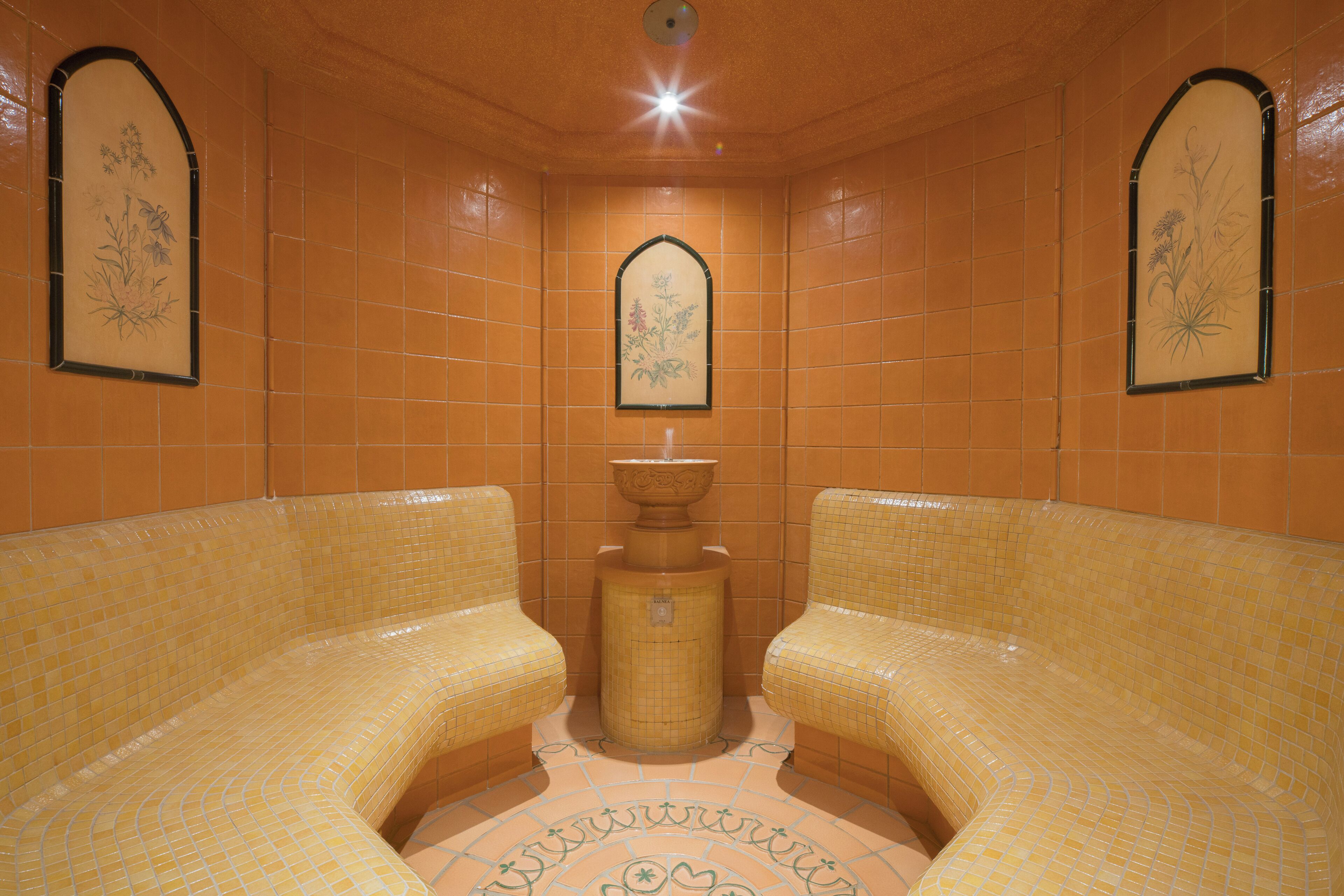 Ein Spa-Raum mit zwei gepolsterten Sitzbereichen, einer zentralen Fontäne und dekorativen Wandfliesen.