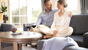 A couple sitting on a grey sofa with a round wooden table between them, containing a plate of grapes and a glass of red wine. - Castello Apartments (Adendorf)
