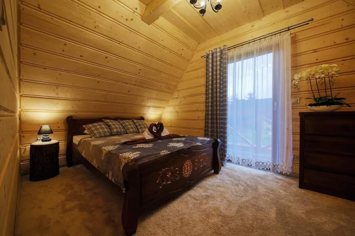 Ein gemütliches Schlafzimmer mit Holzboden, einem Bett mit dekorativem Kopfteil, einem Nachttisch mit Lampe und einem Fenster mit transparenten Vorhängen.