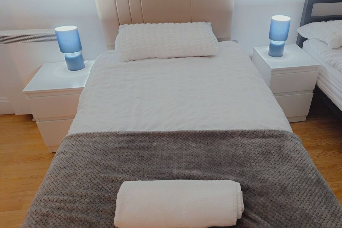 A hotel room with a single bed, two bedside tables with lamps, a grey blanket, and a folded white towel.