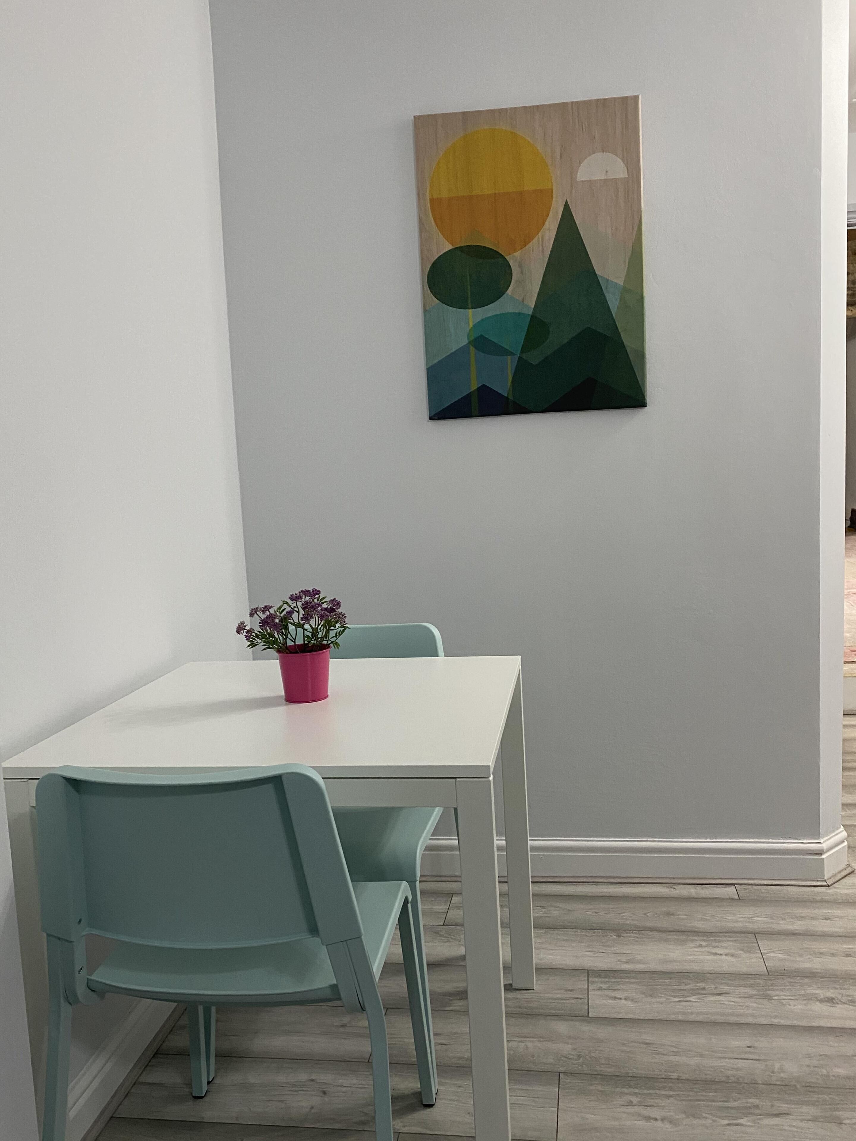 A dining area with a white table and two teal chairs, a pink flower pot, and a geometric abstract painting on the wall.