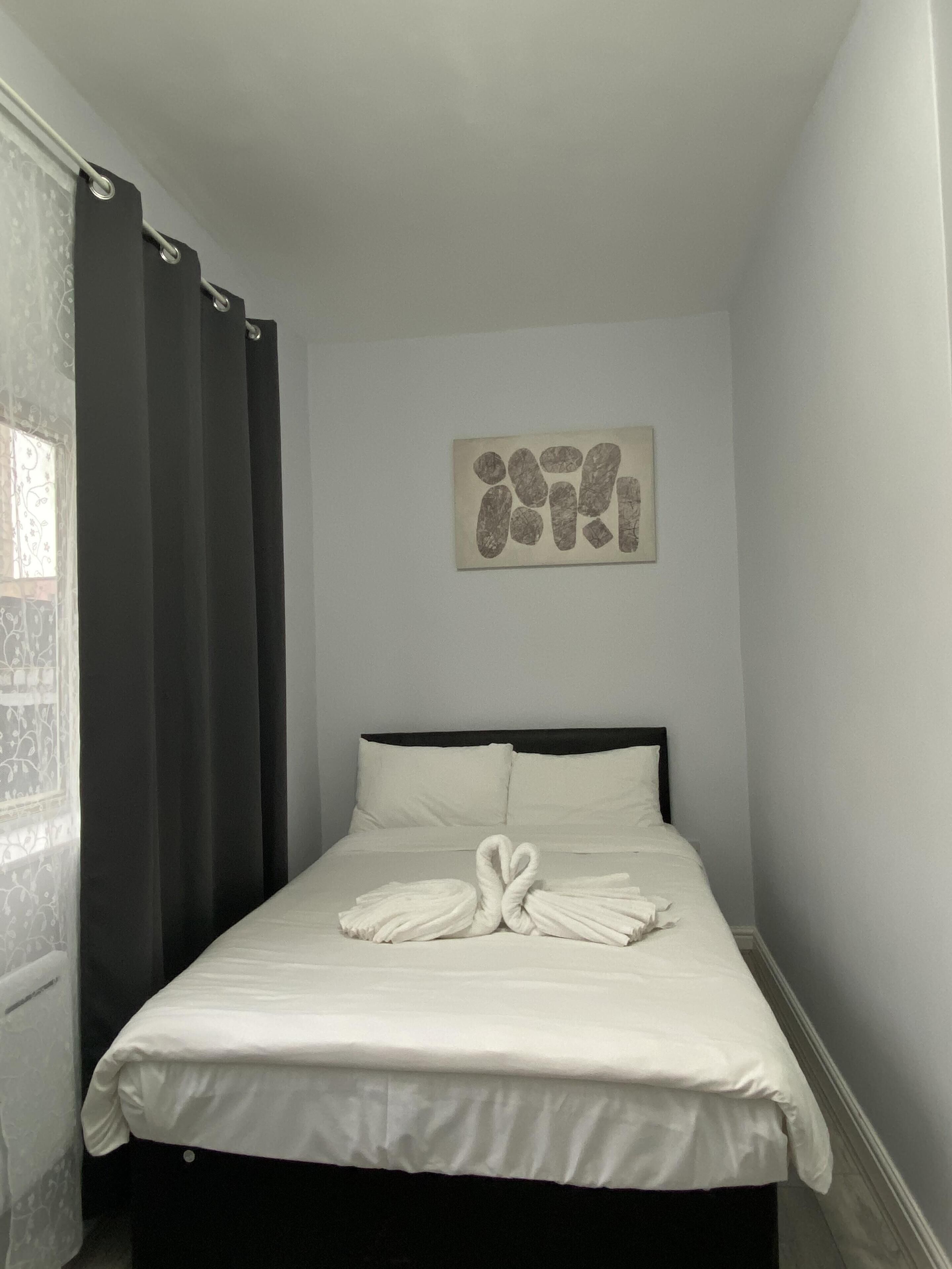Un lit bien fait, avec des serviettes disposées en forme de cœur, un mur sobre orné d’une œuvre d’art abstraite et une fenêtre avec des rideaux à motifs.