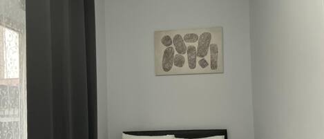 Un lit bien fait, avec des serviettes disposées en forme de cœur, un mur sobre orné d’une œuvre d’art abstraite et une fenêtre avec des rideaux à motifs.