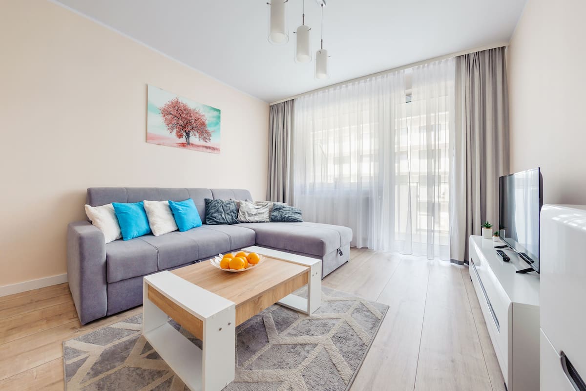 Ein modernes Wohnzimmer mit einem grauen Sofa, einem Couchtisch mit Orangen, einem Fernseher und einem Baumgemälde an der Wand.