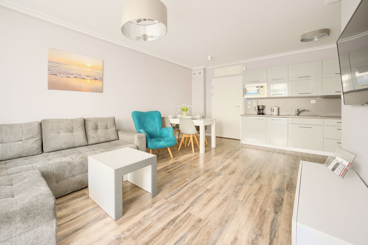 Ein modernes Wohnzimmer mit einem grauen Ecksofa, einem türkisblauen Sessel und einem weißen Couchtisch. An der Wand hängt ein Gemälde von einem Sonnenuntergang, und der Boden ist mit Holz ausgelegt.