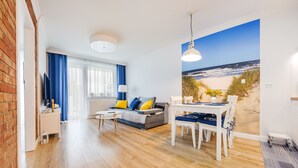 A living room with a sofa, coffee table, and dining table with chairs. There is a large window with a beach view. - Apartamenty Sun & Snow Blizej Morza (Kolobrzeg)
