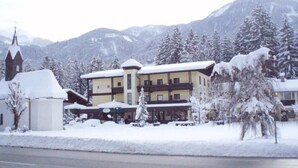 A multi-story hotel with a snow-covered roof and adjacent church, surrounded by snow-laden trees and mountains. - Hotel Vanda (Irschen)