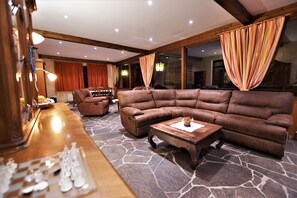 A spacious living area with a large brown leather sectional sofa, a wooden coffee table, and a chessboard on a stone-patterned floor.