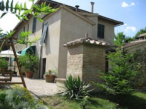 A two-story house with a stone facade, a tiled roof, and a small garden area with potted plants and a wooden bench. - Giannetti bed & breakfast (Montalcino)