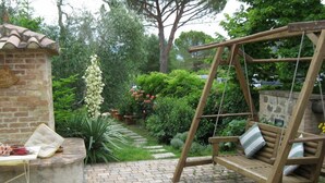 Un patio con una silla colgante, una pequeña construcción de ladrillo y un jardín con diversas plantas y un muro de piedra.