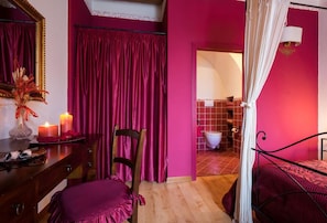 Ein Zimmer mit einem Holzkoffer, einem Sessel, einem Bett mit Baldachin und einem durchgegangenen Tür angedeuteten Badezimmer.
