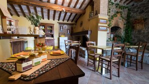 Una cocina rústica con vigas de madera, pared de piedra y chimenea. Hay una zona de comedor con sillas y mesa de madera.