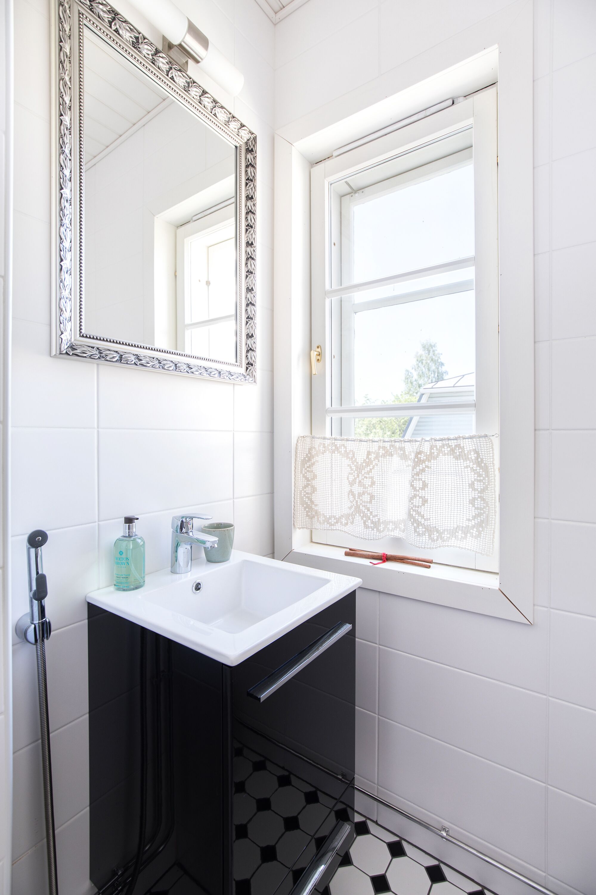 Un baño con un lavabo blanco, un espejo con un marco decorativo, una ventana con cortinas de encaje y un piso de baldosas blanco y negro.
