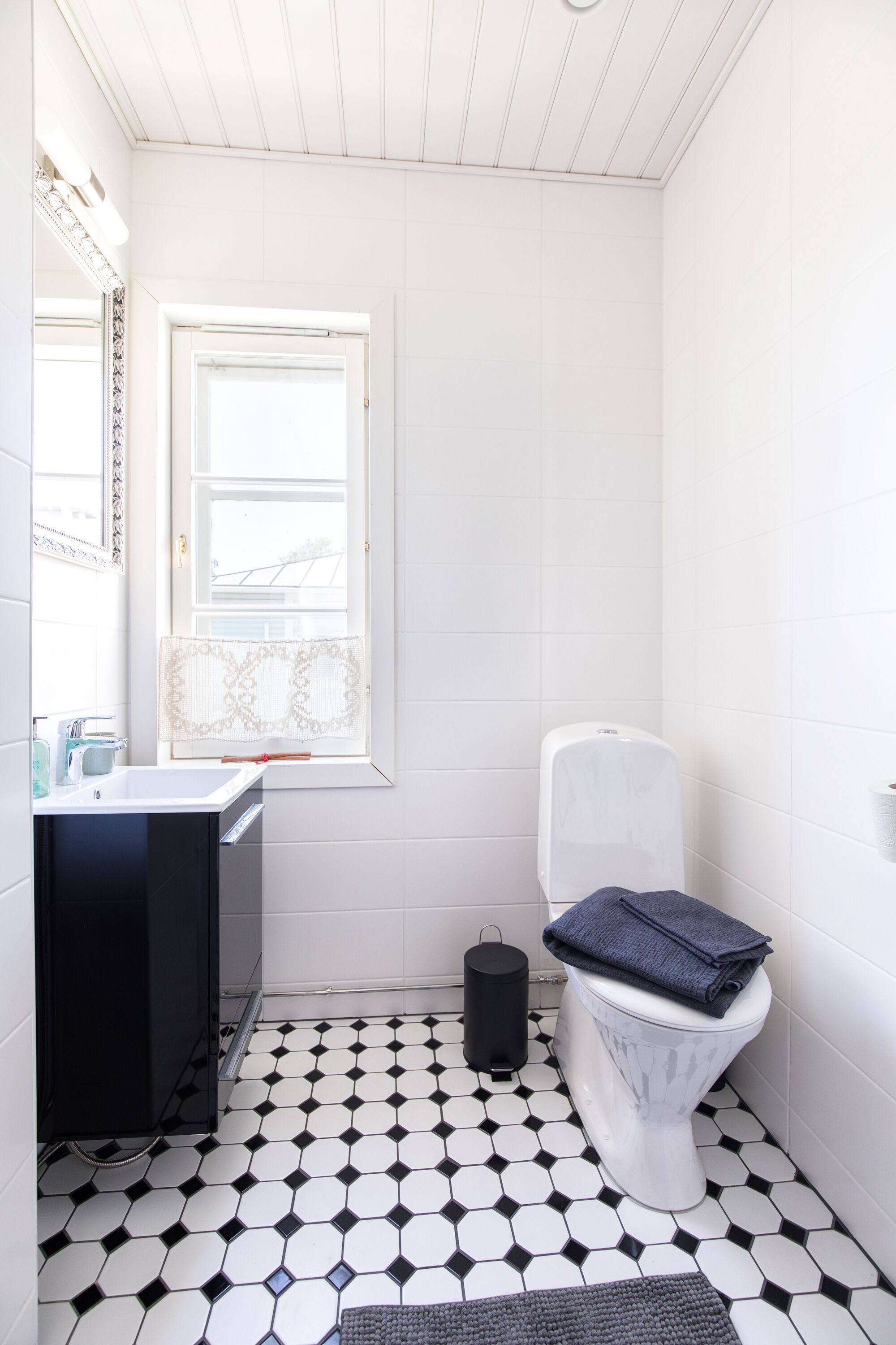A bathroom with a white toilet, a black trash bin, a window with a patterned blind, and a tiled floor.
