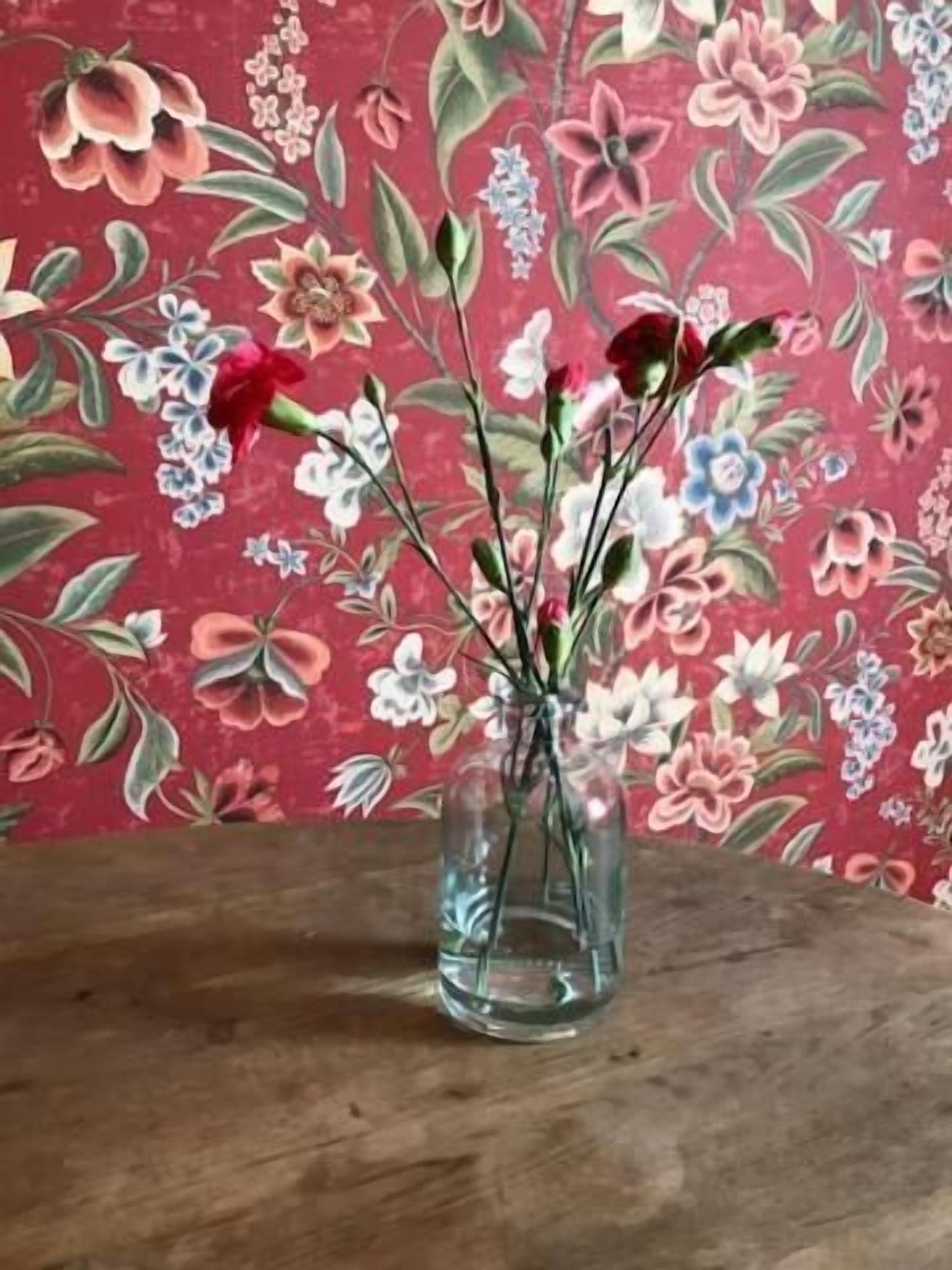 Un vas de vidre transparent amb flors sobre una superfície de fusta davant d'un fons de paper pintat amb motius florals.