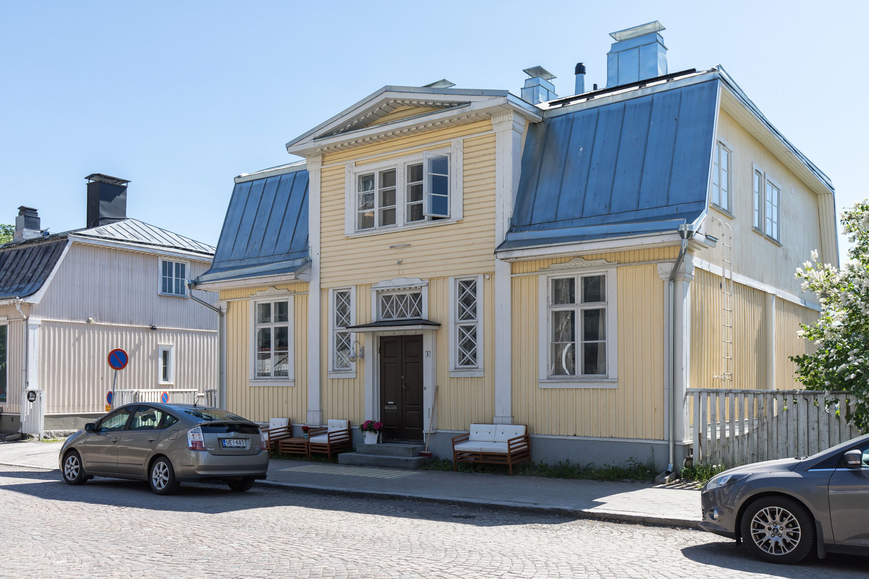 A two-story building with a yellow facade, a dark blue roof, and a front porch with seating.