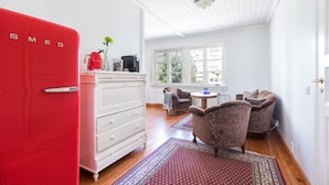 A room with a red refrigerator, white dresser, and patterned rugs. There are two armchairs and a small table with a lamp.
