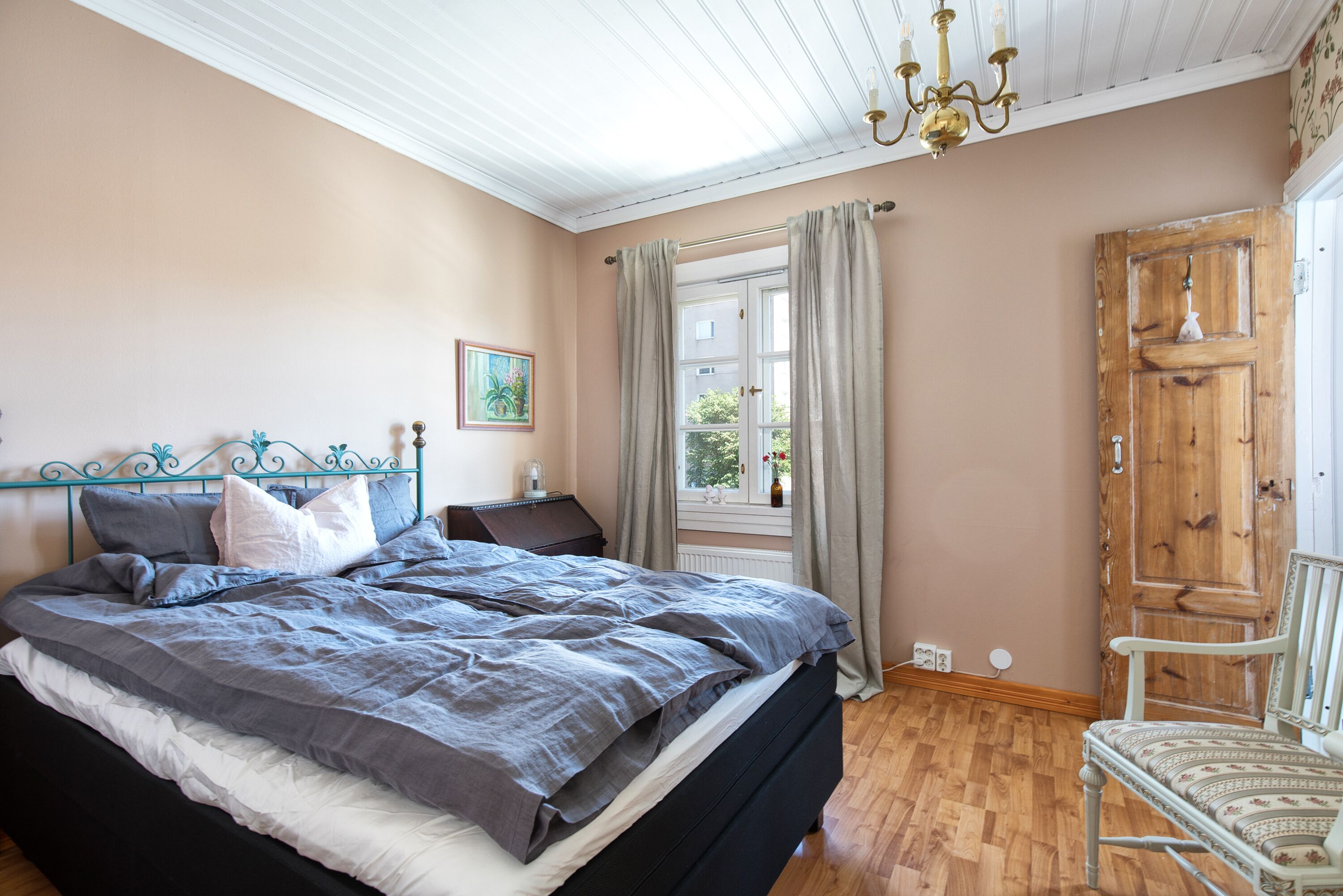A bedroom with a large bed, a wooden wardrobe, a chair, a window with curtains, and a chandelier.