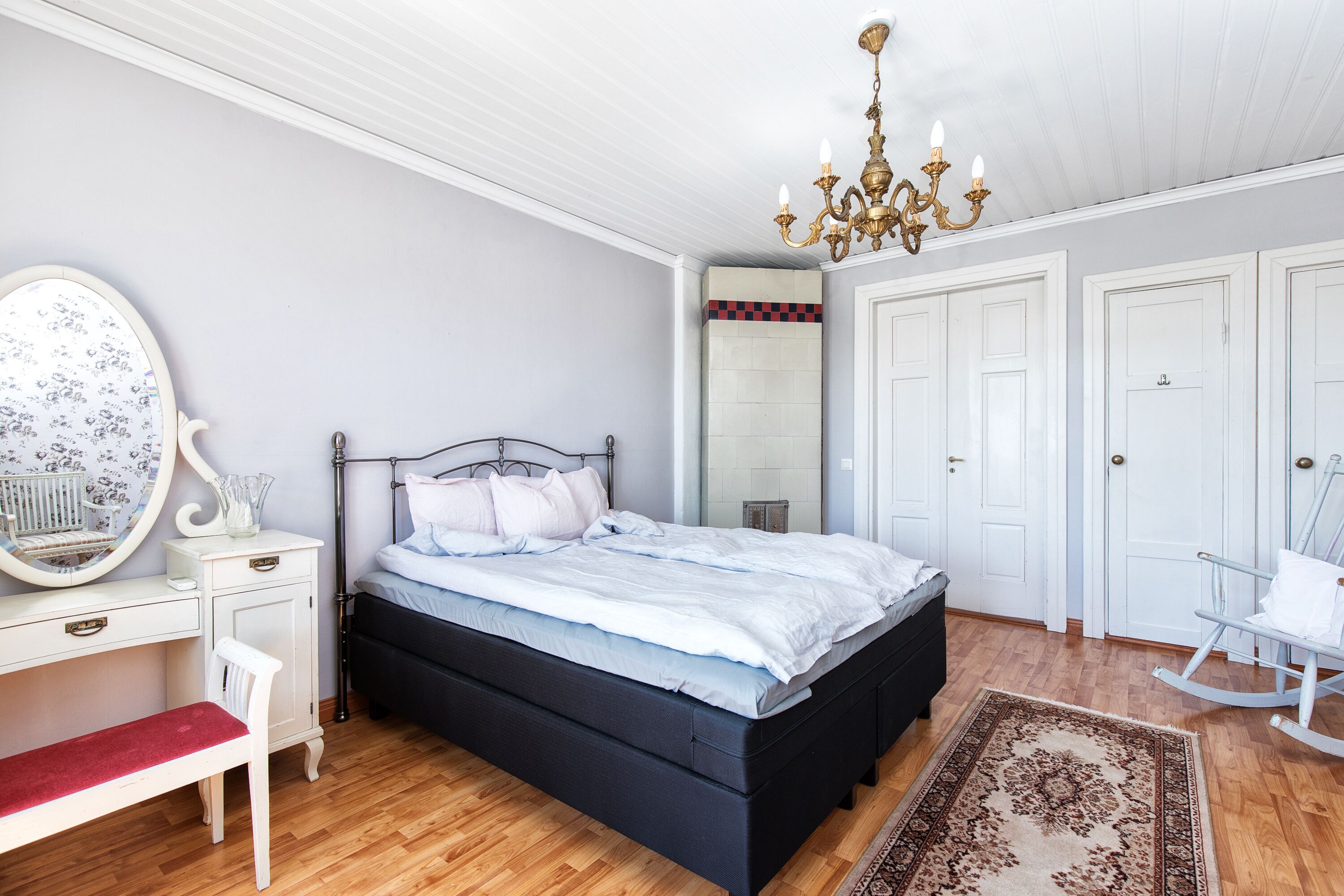 Un dormitorio con una cama, un escritorio, una silla, un sillón mecedora, una araña y un espejo.