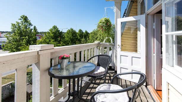 A balcony with a glass table and two chairs, a white door, and a view of trees and buildings.
