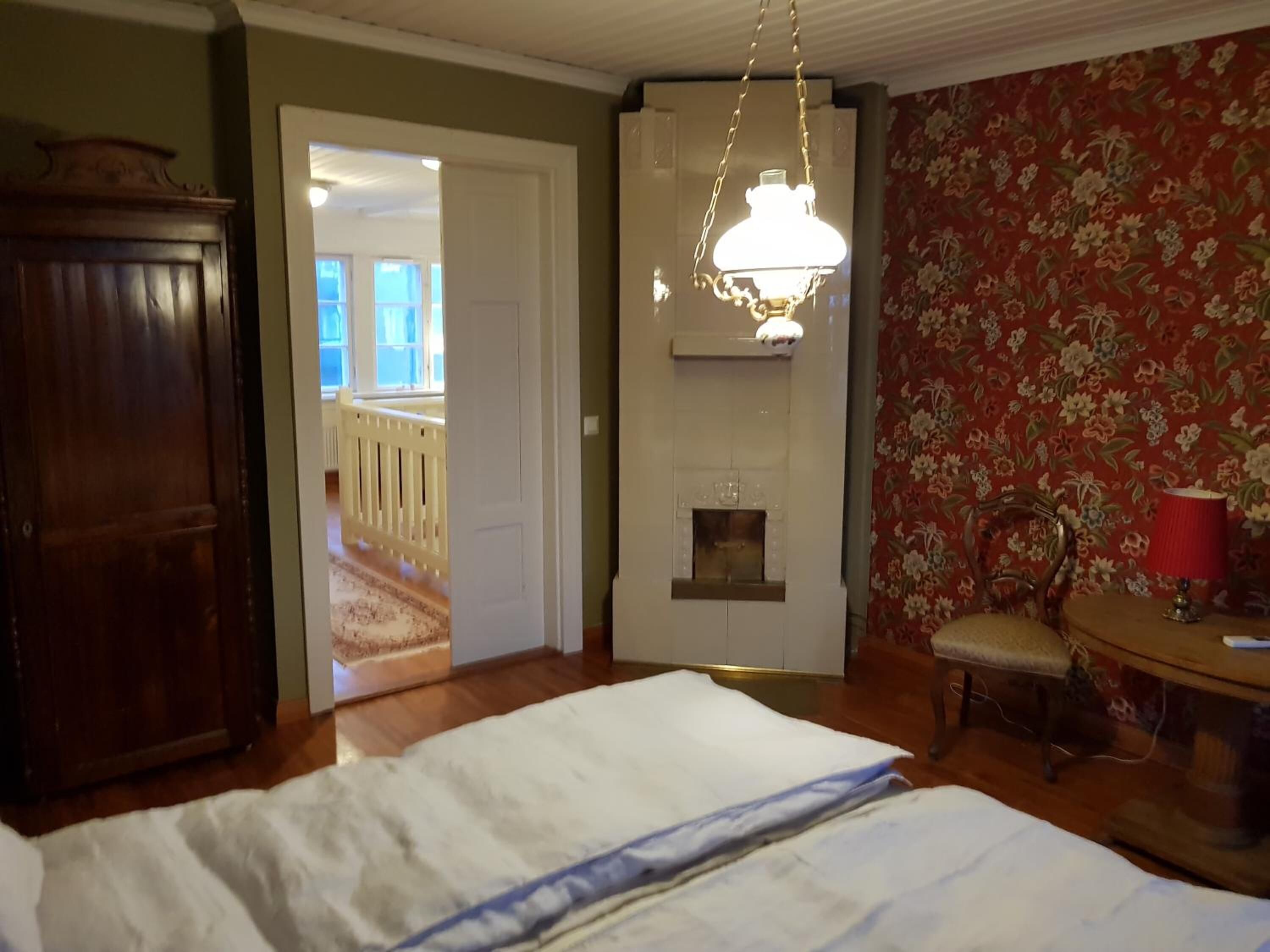 Quarto com cama, guarda-roupa de madeira, lareira, poltrona e mesa com abajur.