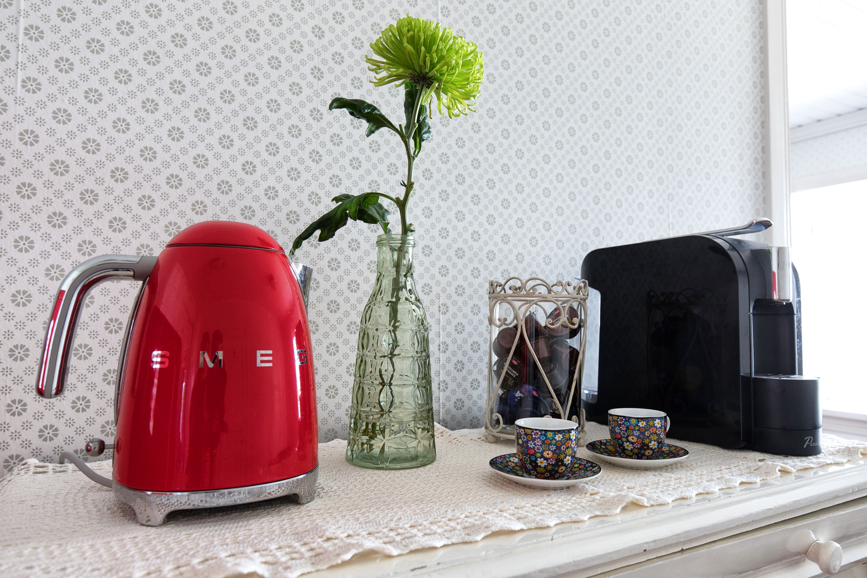A kitchen counter with a red electric kettle, a glass vase with a green flower, a coffee maker, and a decorative metal container with a cup and saucer.