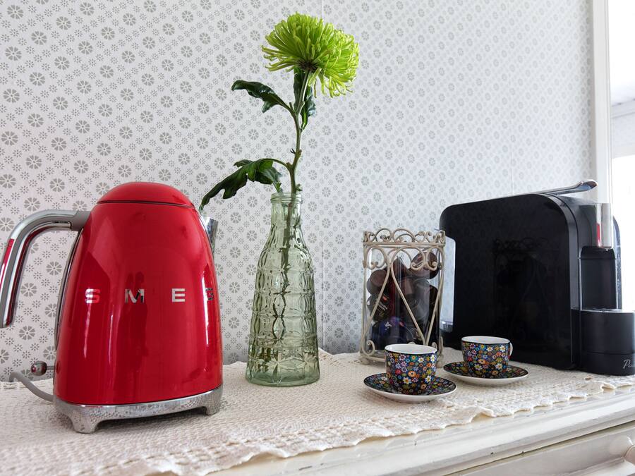 En køkkenbordplade med en rød elkedel, en glasskål med en grøn blomst, en kaffemaskine og en dekorativ metalbeholder med kop og underkop.