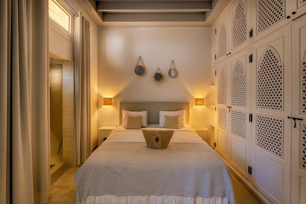 A hotel room with a large bed, two bedside tables, a nightstand, a wall-mounted mirror, and a wardrobe with intricate designs.