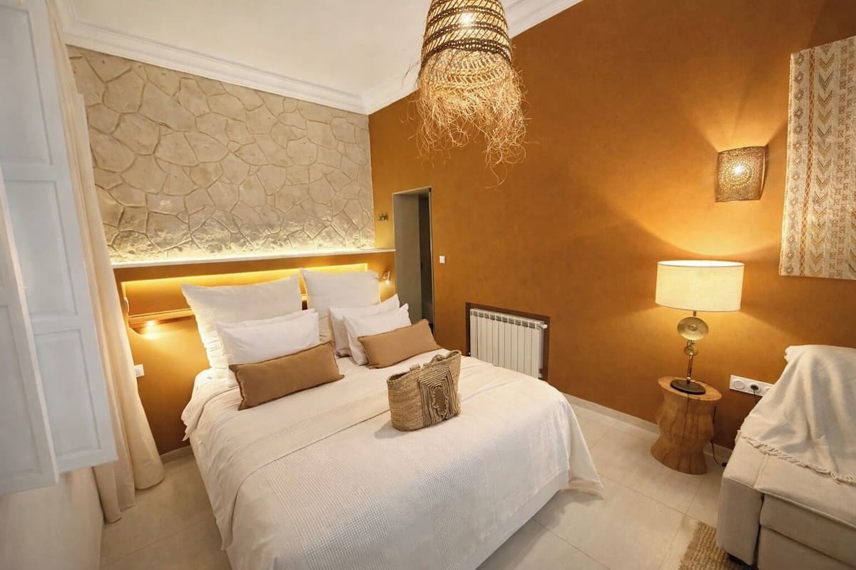 A hotel room with a stone accent wall, a bed with white linens and brown pillows, a wooden nightstand with a lamp, and a radiator.