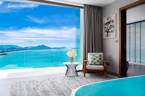 A modern hotel room with a large balcony, a view of the sea, a pool, and a chair with a cushion.