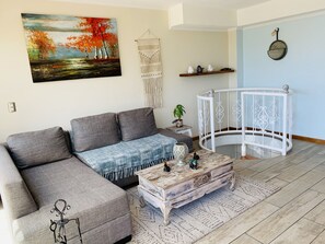 A living room with a grey sectional sofa, a rustic coffee table, a decorative wall hanging, and a white spiral staircase.