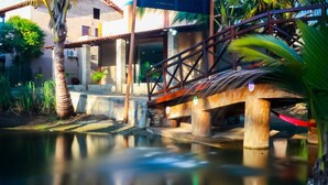 A wooden bridge over a calm water body, with a building and palm trees in the background. - Pousada Recanto do Mar (Camocim)