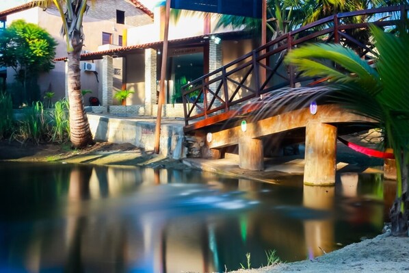 A wooden bridge over a calm water body, with a building and palm trees in the background.