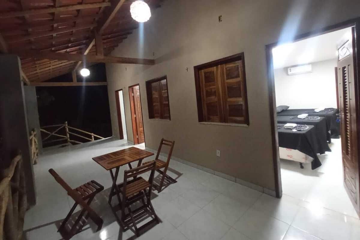 Una habitación con una mesa y sillas de madera, un ventilador de techo y una puerta que da a otra habitación con camas.