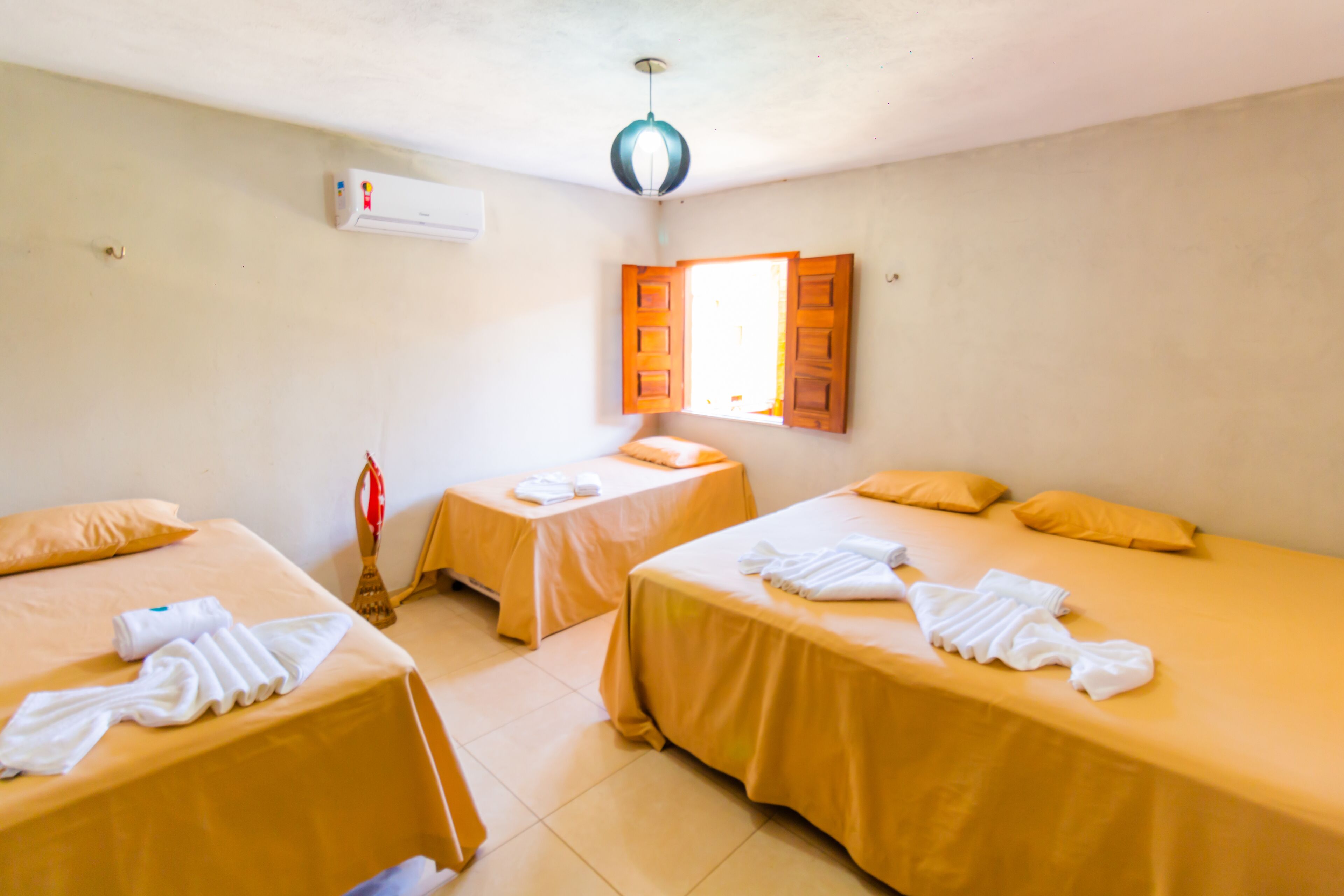 Habitación de hotel con dos camas individuales, cada una con una colcha amarilla y toallas blancas. Se ve una ventana con contraventanas de madera, un equipo de aire acondicionado en la pared y una lámpara en el techo.
