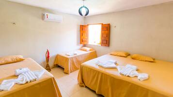 Habitación de hotel con dos camas individuales, cada una con una colcha amarilla y toallas blancas. Se ve una ventana con contraventanas de madera, un equipo de aire acondicionado en la pared y una lámpara en el techo.