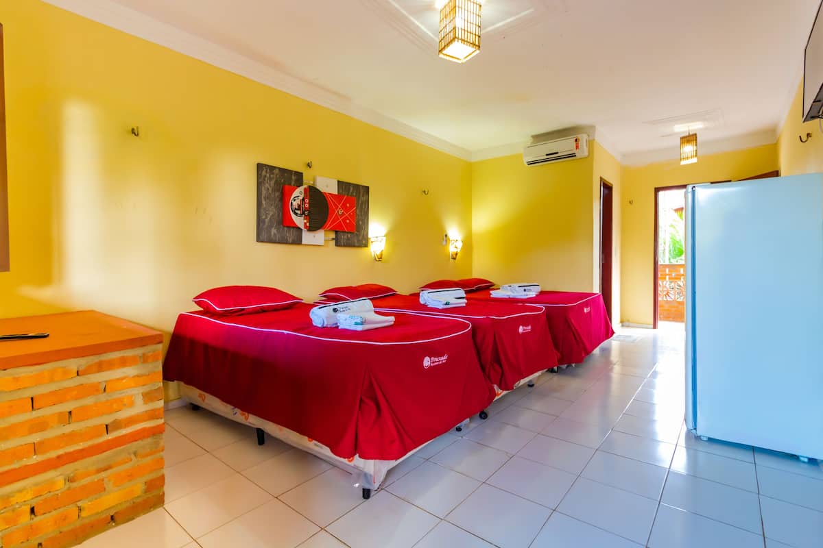 Habitación de hotel con cuatro camas cubiertas con tela roja, pared de ladrillo, techo blanco y refrigerador.