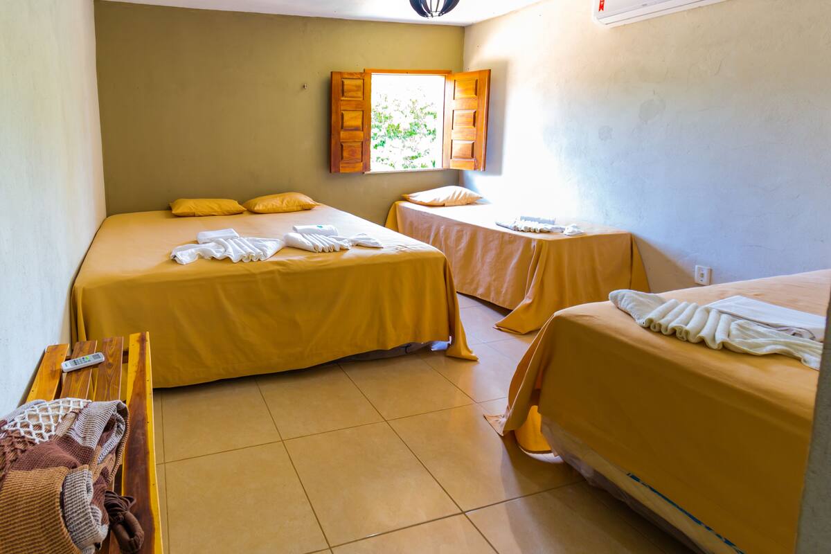 Habitación con dos camas, cada una con cubrecama y almohadas amarillas, una mesa de madera con un control remoto y una ventana con contraventanas de madera.